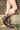 Outdoors by a brick and concrete wall, a person wears AZALEA WANG IMOGINE Brown Multi Folded Detail Stiletto Boots with pointed toes, faux leather that fades from dark brown to beige, and metallic stud accents.