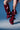 A person wears the AZALEA WANG IMOGINE BURGUNDY STILETTO SNAP BOOT—oversized, sculptural deep red leather with folds and metallic studs—against a blue background, highlighting the bold pointed toe stiletto style. Only the legs are visible.