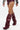 A person wears high-waisted maroon shorts, a long-sleeve top with sleeve text, and AZALEA WANG IMOGINE BURGUNDY STILETTO SNAP BOOT—pointed toe, faux leather, knee-high stilettos—while standing against a plain white background.