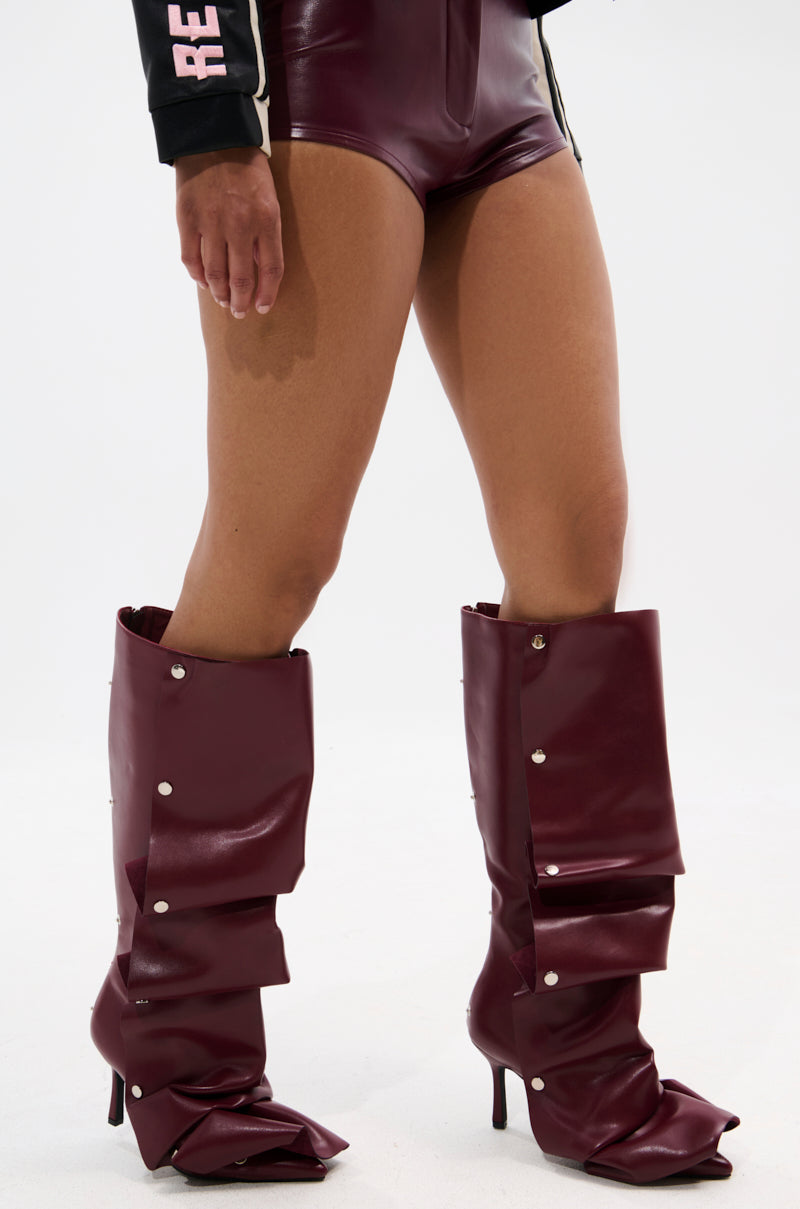 Wearing high-waisted maroon leather shorts, a black sleeve with white and pink text, and the AZALEA WANG IMOGINE BURGUNDY STILETTO SNAP BOOT with pointed toes and silver buttons, a person stands against a white background.