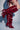 A person wears the AZALEA WANG IMOGINE BURGUNDY STILETTO SNAP BOOT, featuring ruffled details, metallic studs, and red thigh straps that accentuate these bold high-heeled, over-the-knee pointed toe boots.