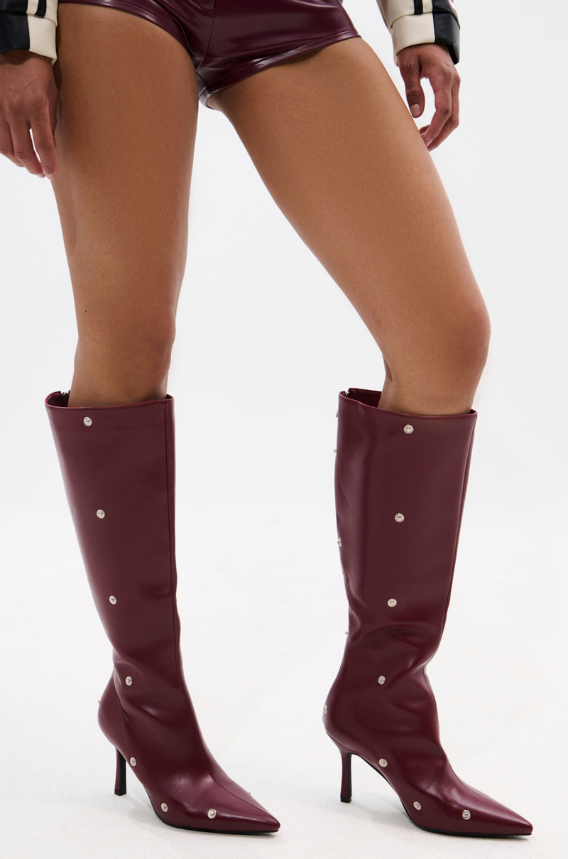 Wearing high-waisted dark purple vinyl shorts and AZALEA WANG IMOGINE BURGUNDY STILETTO SNAP BOOTS, a person stands against a plain white background.