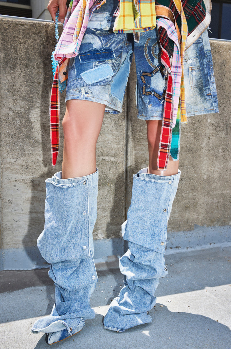 Standing on a concrete surface before a weathered wall, a person wears patchwork denim shorts, mixed plaid shirts tied at the waist, and AZALEA WANG IMOGINE DENIM SNAP BOOTS with an asymmetrical shaft.