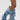 Person wearing the AZALEA WANG IMOGINE DENIM SNAP BOOT—knee-high, pointed toe, stiletto heel, crafted from slouchy blue denim with metal snap details—stands against a plain white background.