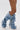 Person wearing the AZALEA WANG IMOGINE DENIM SNAP BOOT—knee-high, pointed toe, stiletto heel, crafted from slouchy blue denim with metal snap details—stands against a plain white background.