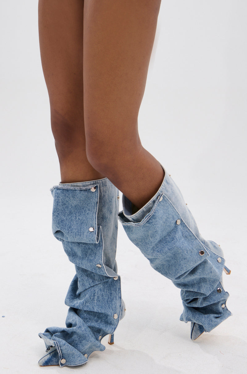 A person wearing the AZALEA WANG IMOGINE DENIM SNAP BOOT, featuring silver buttons, a pointed toe, and stiletto heel, crosses their bare legs against a plain white background.
