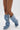 A person wearing the AZALEA WANG IMOGINE DENIM SNAP BOOT, featuring silver buttons, a pointed toe, and stiletto heel, crosses their bare legs against a plain white background.