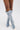 A person with crossed legs wears the AZALEA WANG IMOGINE DENIM SNAP BOOT—light blue, knee-high denim boots with a pointed toe, metallic studs, and a sleek stiletto heel—standing on a white background.