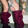 A person in a floral embroidered jacket and denim shorts adjusts AZALEA WANG IMOGINE PURPLE OMBRE FOLDED DETAIL STILETTO BOOTS while standing indoors on a textured floor in front of glass blocks.