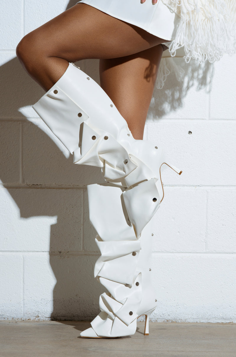 Legs wearing the AZALEA WANG IMOGINE WHITE BOOT, over-the-knee high-heeled boots with metallic studs, stand against a white brick wall. These faux leather boots have a slouchy, textured design and stiletto heel, paired with the lower portion of a white fringed outfit.