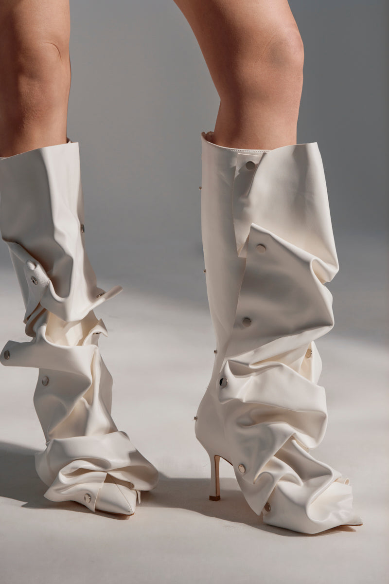 A close-up of a person wearing the AZALEA WANG IMOGINE WHITE BOOT, featuring cream-colored, ruched faux leather and visible buttons that create a sculptural, oversized look against a softly lit neutral background.