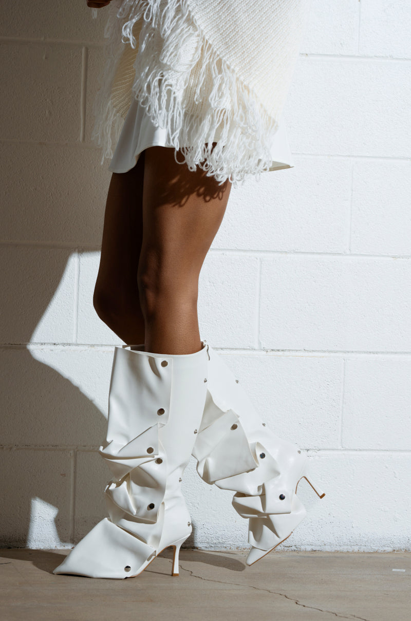 The image features the lower half of a person wearing AZALEA WANG IMOGINE WHITE BOOT, knee-high and adorned with buttons, paired with a fringed faux leather outfit. They stand against a white brick wall on a concrete floor.