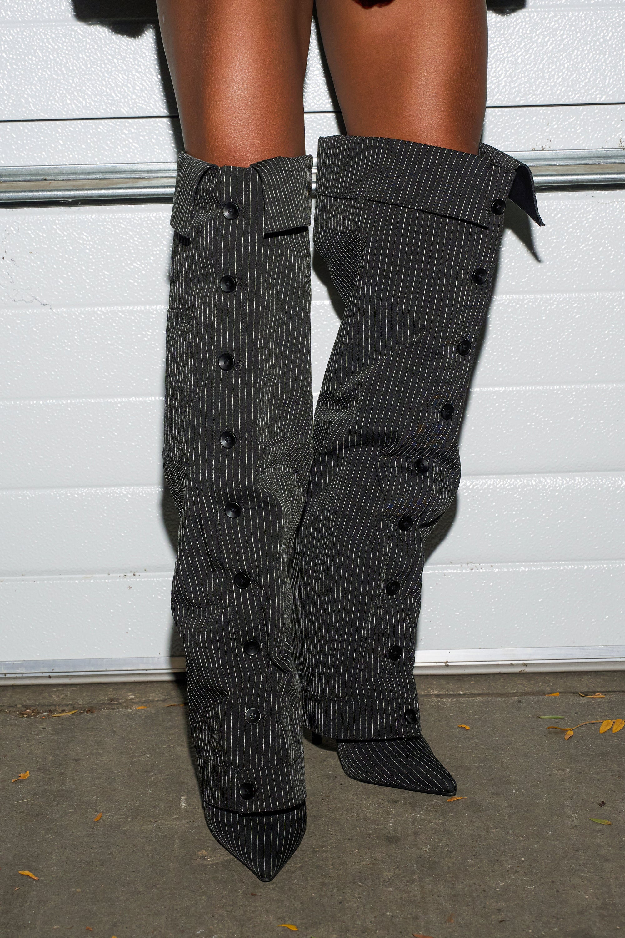 A person wears the AZALEA WANG IMRI BLACK SHIRTING STRIPED STILETTO BOOT with pointed toes, standing on concrete before a white wall.