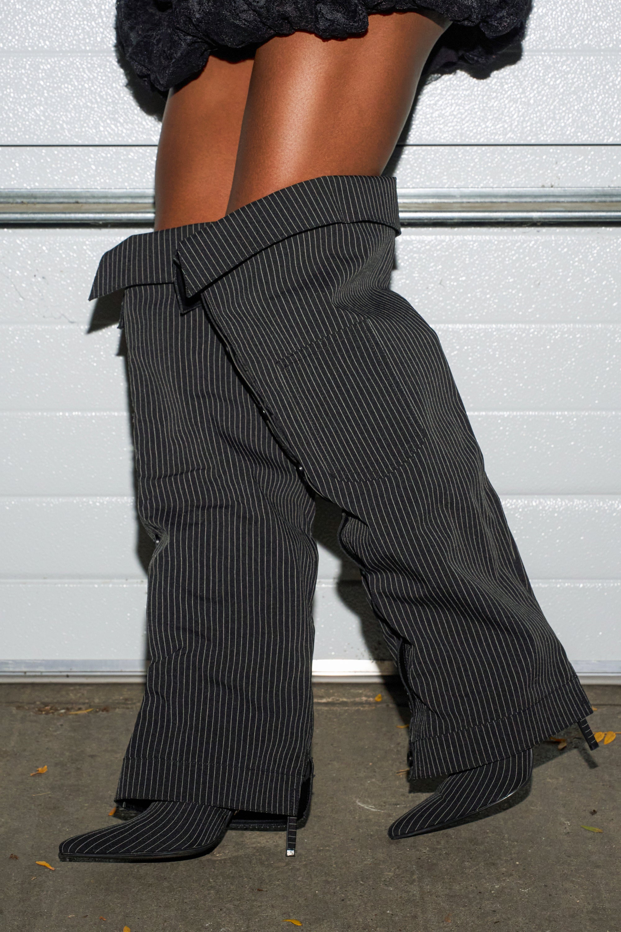 Someone wears AZALEA WANG IMRI BLACK SHIRTING STRIPED STILETTO BOOTS, styled to resemble oversized suit pants, paired with a black textured skirt, standing on concrete before a white garage door.