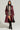 A model stands against a plain background wearing a burgundy leather trench coat over the FORGET ABOUT ME MINI DRESS, which features a black-and-white colorblock chevron pattern, paired with fishnet stockings. They accessorize with black boots, a black hood, glasses, and a black handbag.