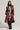 A person stands confidently in a stylish outfit. They wear a maroon leather trench coat over an AKIRA Label FORGET ABOUT ME MINI DRESS with a colorblock chevron pattern, black knee-high boots, and a black headscarf. They also wear brown glasses and hold a black leather bag.