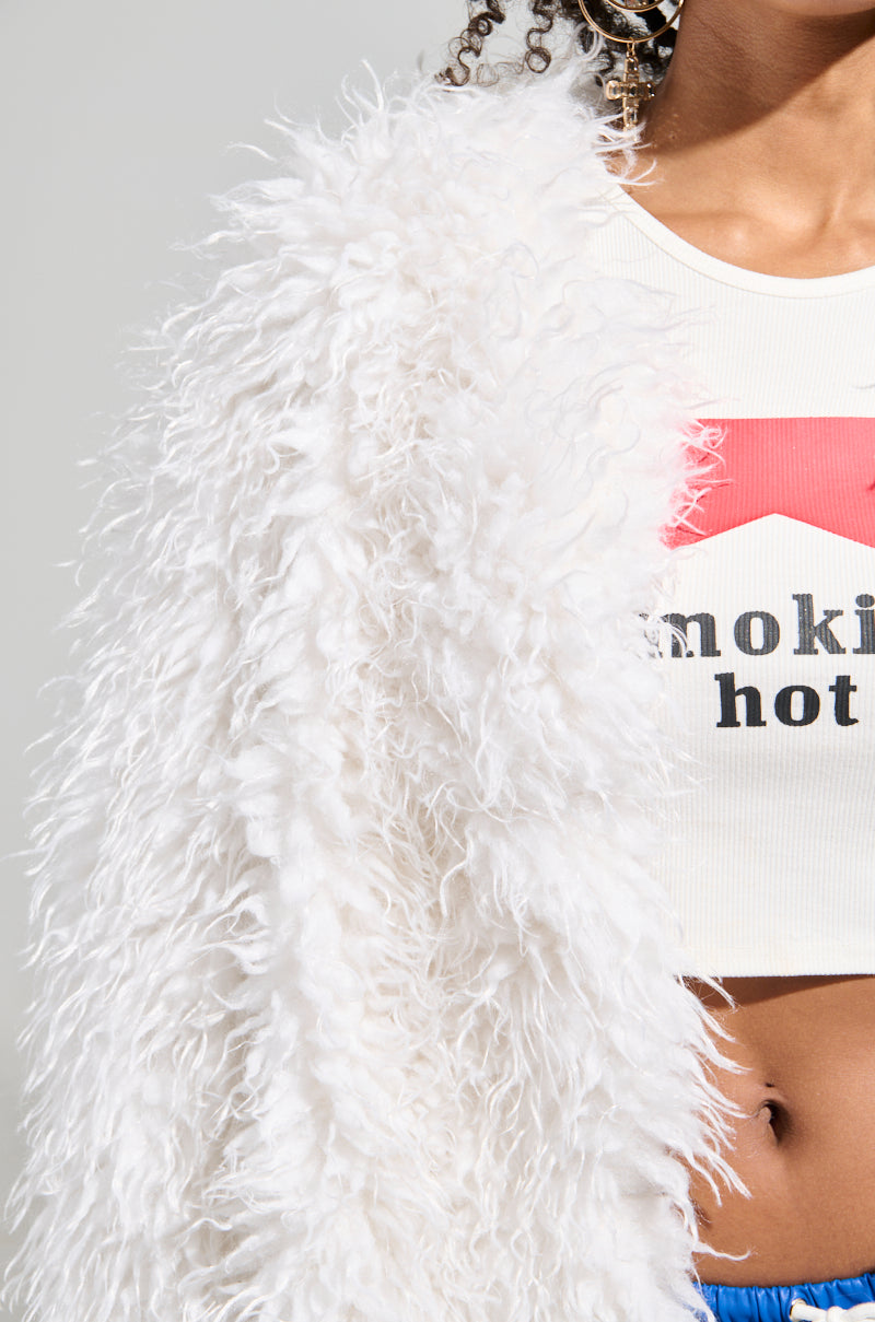 The individual wears an INA Mohair Fur Trench in a fluffy white texture over a cropped white top that reveals the partial text, "smoking hot." The neutral background highlights the luxurious textures and colors of the outerwear.