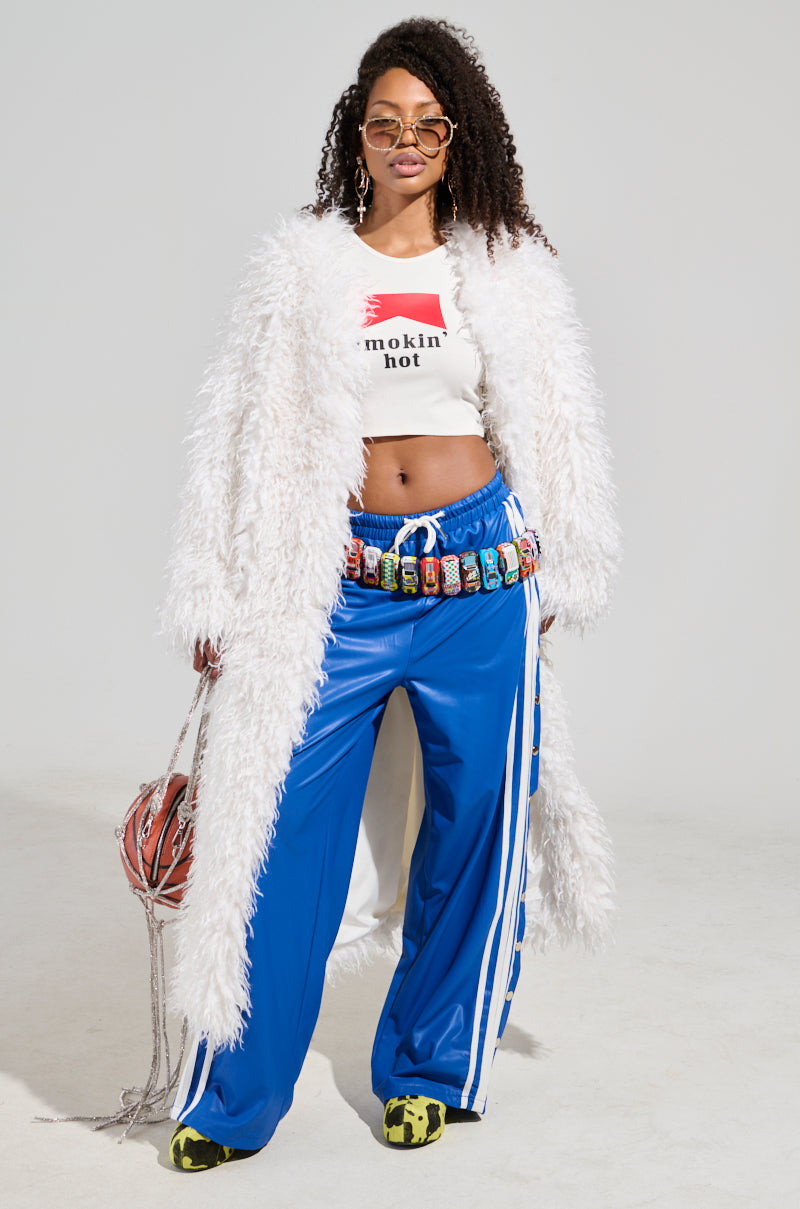 A woman stands confidently wearing a luxurious outerwear piece—an INA MOHAIR FUR TRENCH—and a shirt with "smokin' hot" emblazoned on it. She pairs these with blue track pants and green sneakers while accessorizing with unique sunglasses and holds a netted basketball.