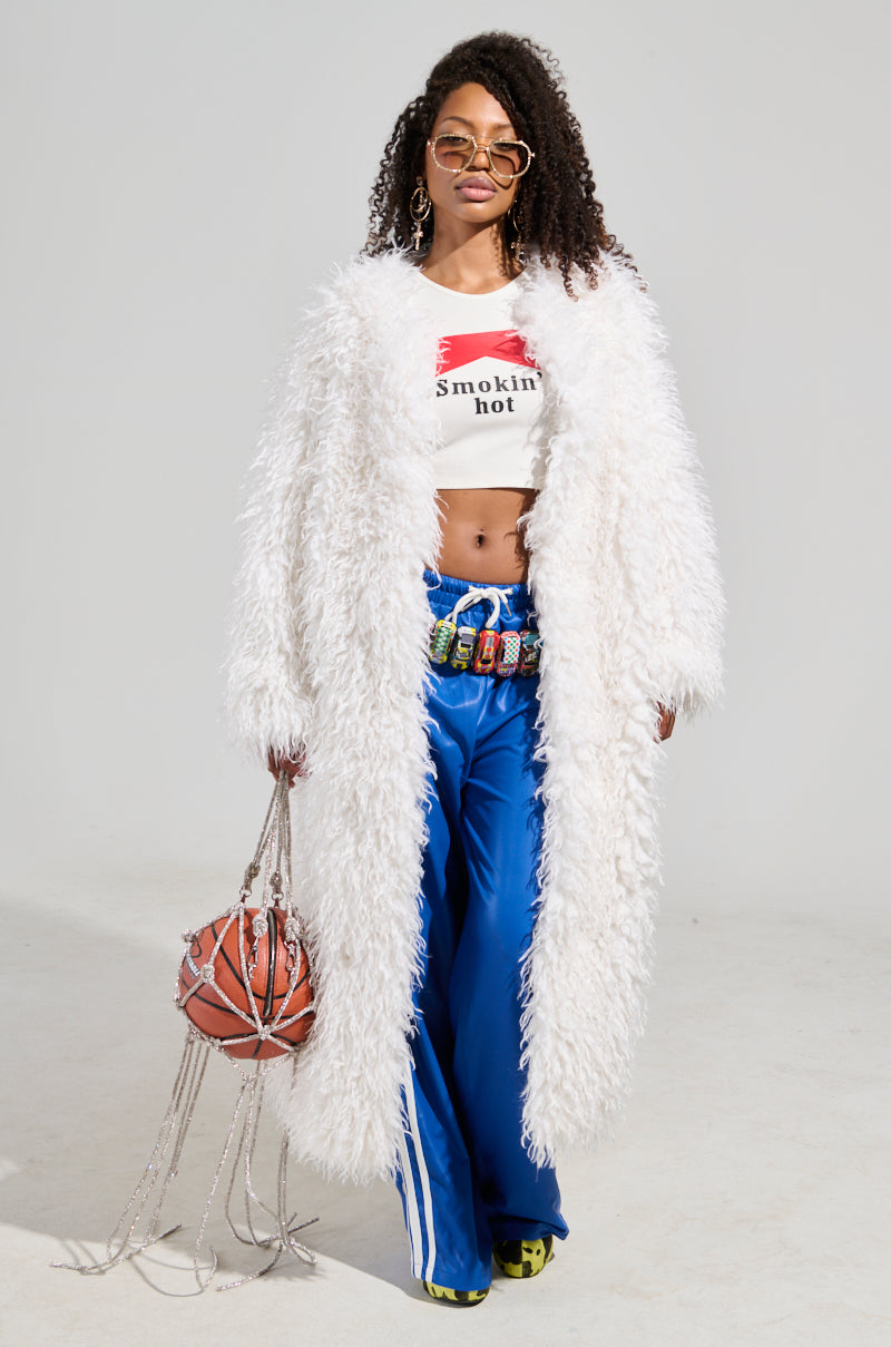 Wearing the INA MOHAIR FUR TRENCH, a person exudes luxury with their fluffy white coat and blue track pants. They hold a basketball embellished with sparkling chains and sport a crop top with text, accentuated by chic sunglasses and a vibrant belt, embodying an effortlessly stylish look.