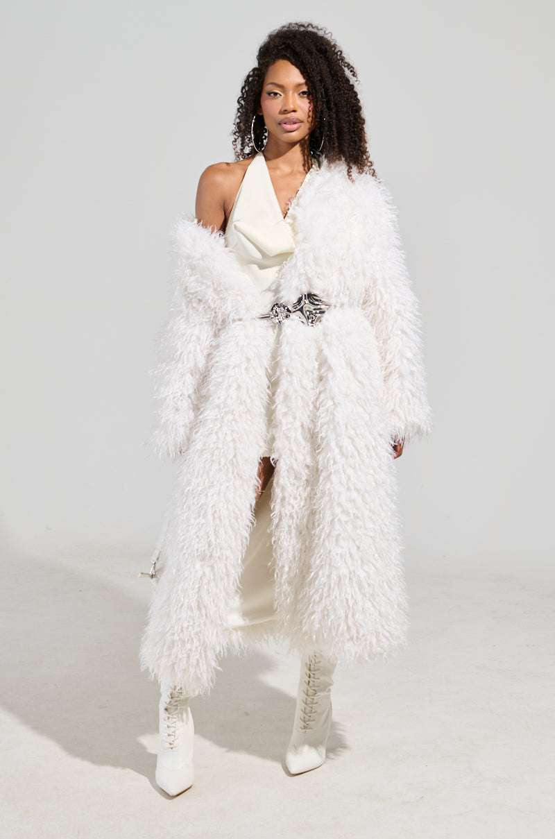 A person models the INA MOHAIR FUR TRENCH over a chic white outfit with matching heeled boots, posed against a plain background. This classic and stylish ensemble features asymmetrical elements and textured layers, highlighting the luxurious outerwear and elegance of the longline silhouette.