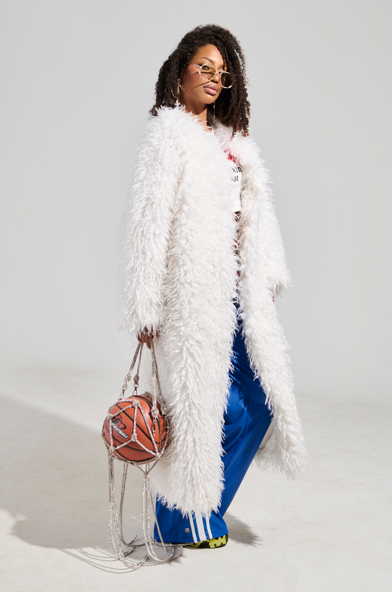 Dressed in the luxurious INA MOHAIR FUR TRENCH over a graphic shirt and blue pants, the individual confidently holds a basketball-shaped bag. With curly hair and sunglasses, they strike a confident pose against a plain backdrop.