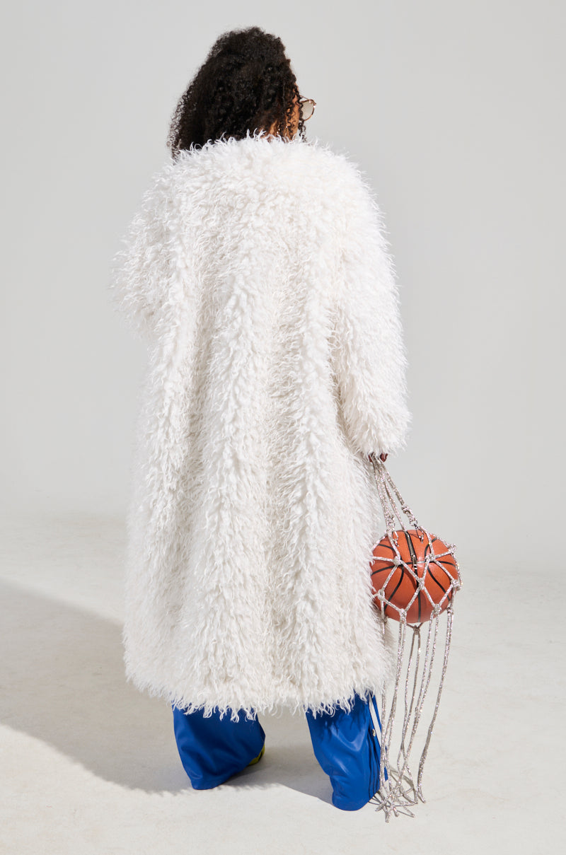 A person with curly hair is elegantly draped in an INA MOHAIR FUR TRENCH—a luxurious long, fluffy white coat—and blue pants. They hold a basketball in a knotted rope bag, all set against a simple, neutral background.