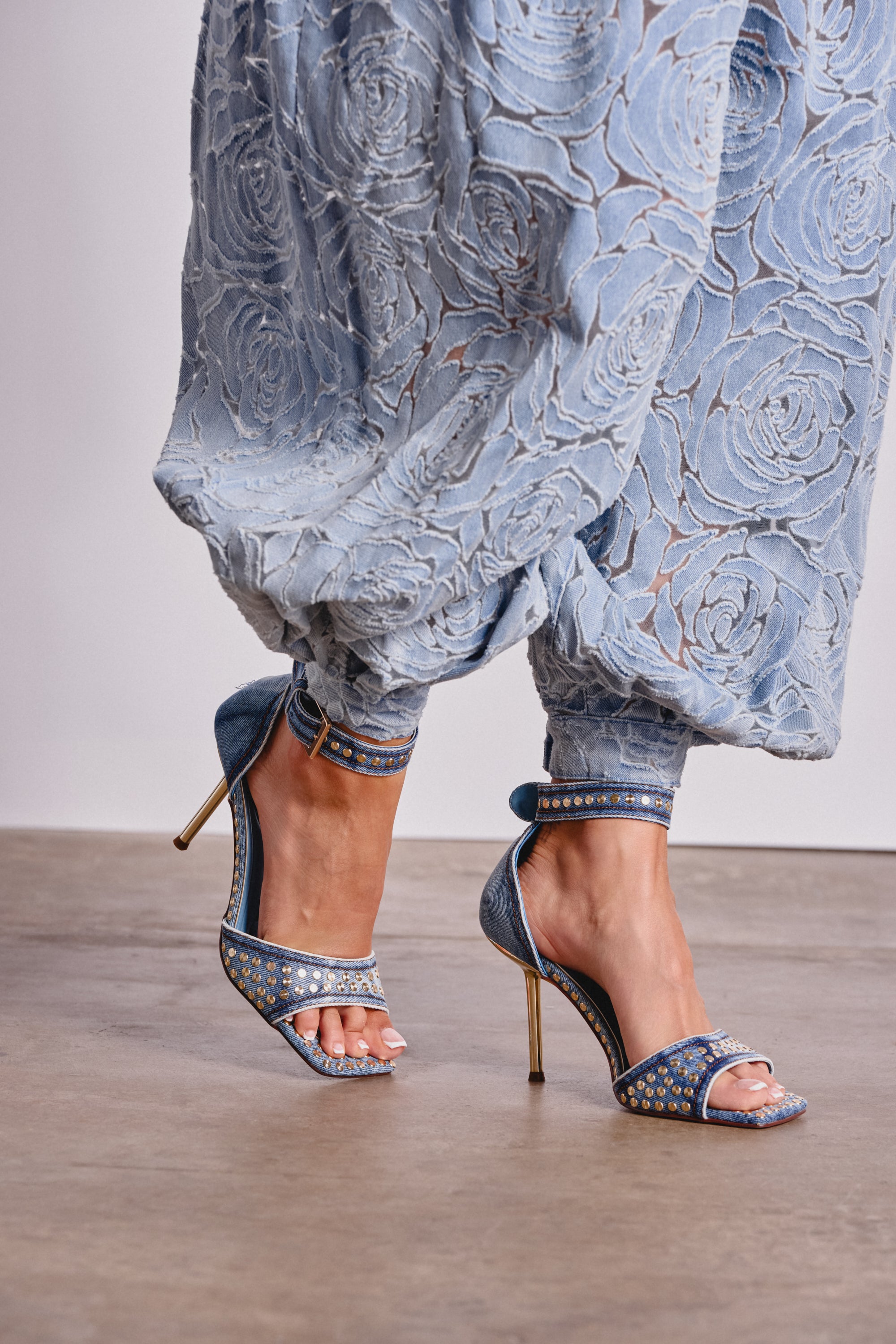 A person stands on a brown floor wearing light blue, floral-patterned, loose-fitting pants and AKIRA GOLD INANA DENIM STUD EMBELLISHED STILETTO SANDALS. 0