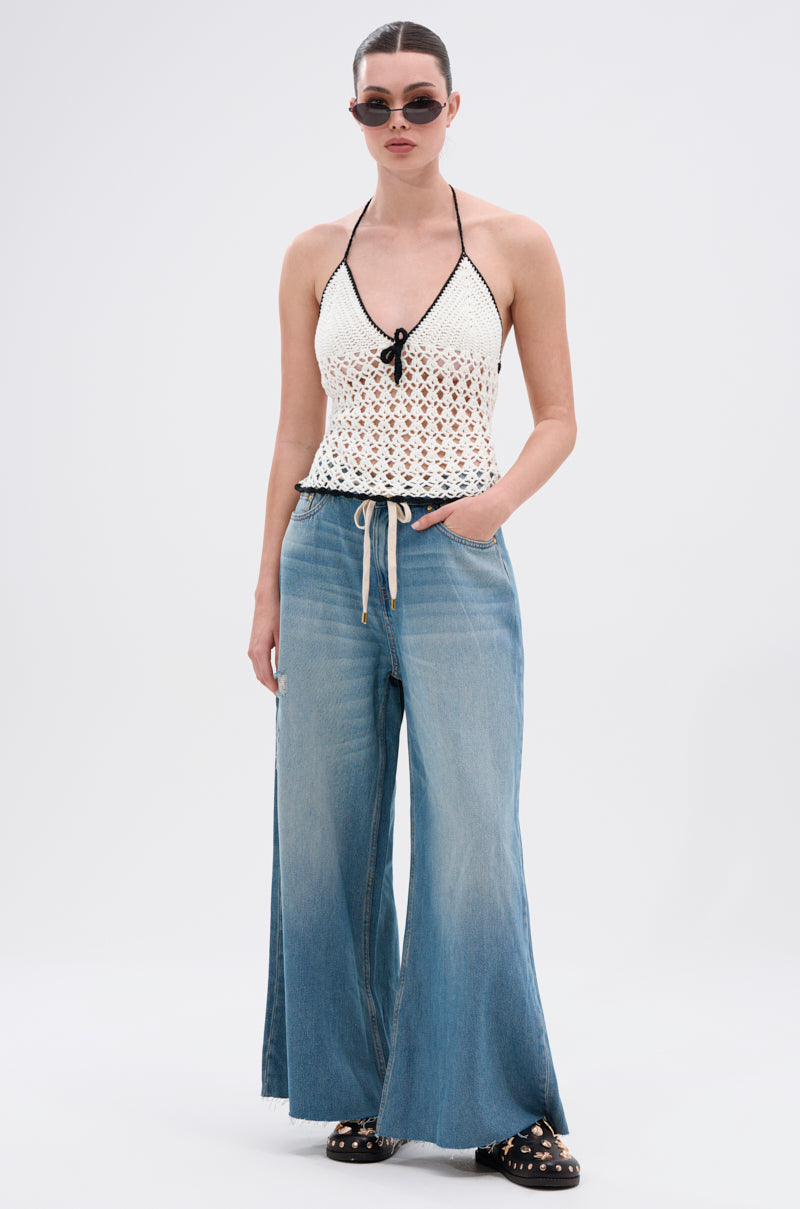 A woman in sunglasses and a white crochet halter top pairs INDIRON JEANS—wide-leg, vintage-wash denim—with black platform sandals featuring gold studs, standing against a plain white background.