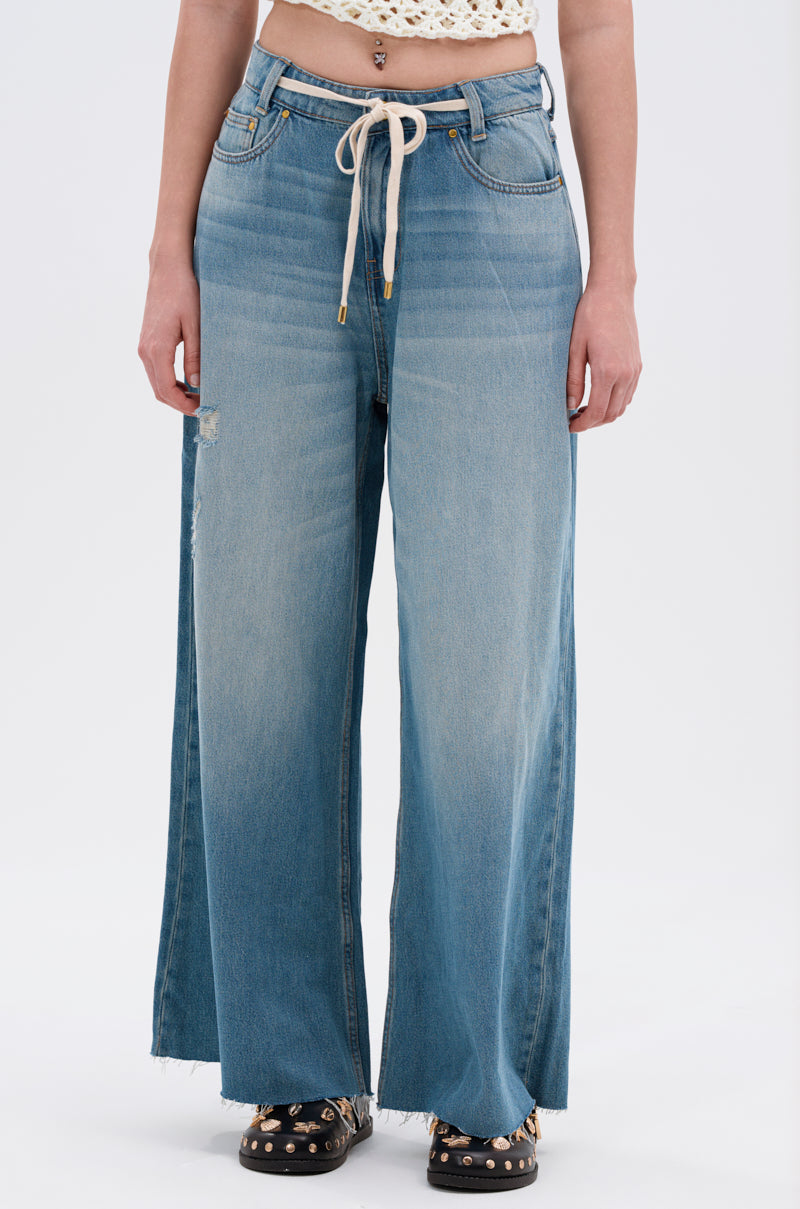 Someone wearing INDIRON JEANS with a wide-leg silhouette in vintage-wash denim, drawstring waist, and a distressed patch above one knee, styled with a white crocheted crop top and black studded sandals.