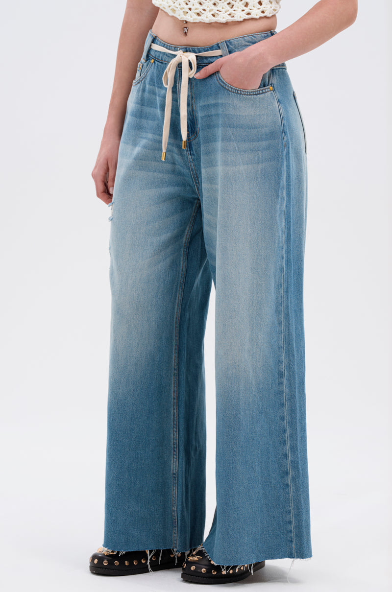 A person wears high-waisted INDIRON JEANS with a wide-leg silhouette and drawstring waist, paired with a crocheted crop top and black studded shoes. The cropped photo shows them from shoulders down against a plain background.