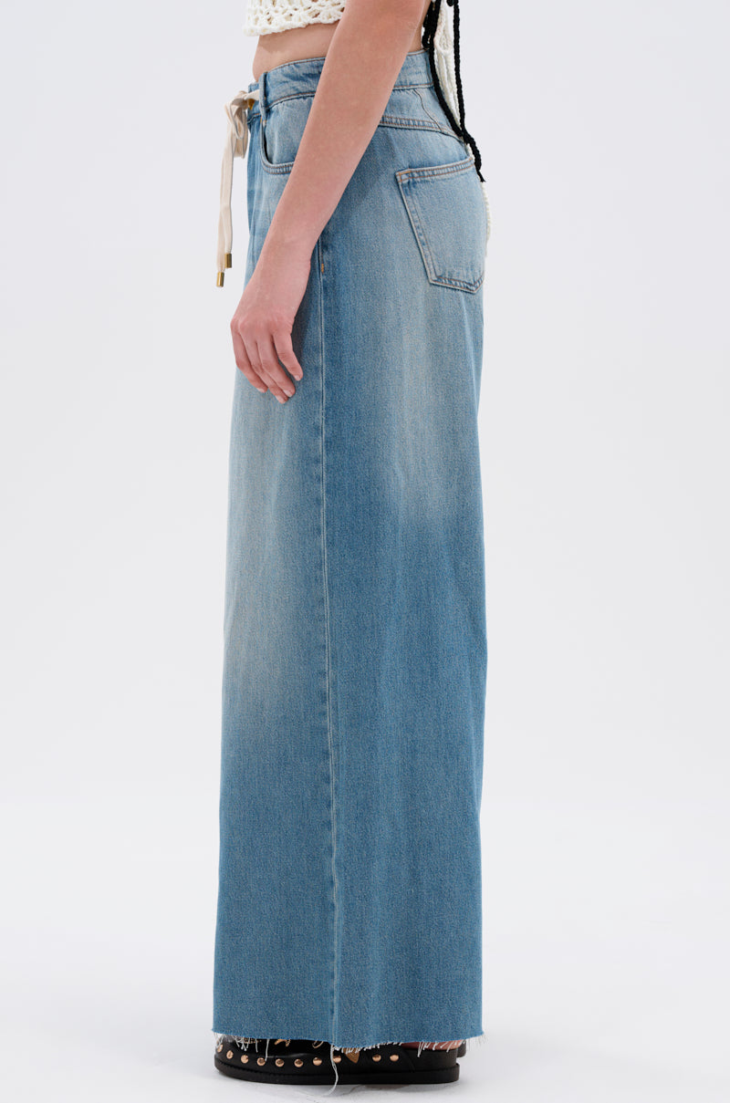 A person in profile wears INDIRON JEANS—vintage-wash, high-waisted, wide-leg jeans with a raw hem and back pocket—styled with black shoes and a crocheted top against a plain white background.