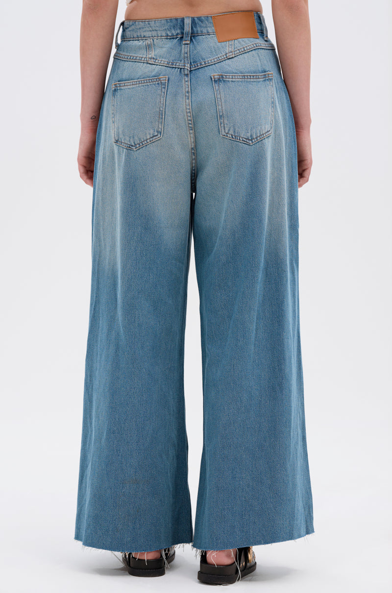 A person is seen from behind wearing INDIRON JEANS in light blue vintage-wash denim with a wide-leg fit, frayed hems, two back pockets, and a brown waistband patch, paired with black shoes.