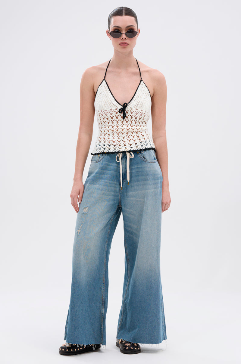 Wearing sunglasses, a white crochet halter top, INDIRON JEANS with a wide-leg silhouette and drawstring waist, and black studded shoes, a person stands against a plain light background.