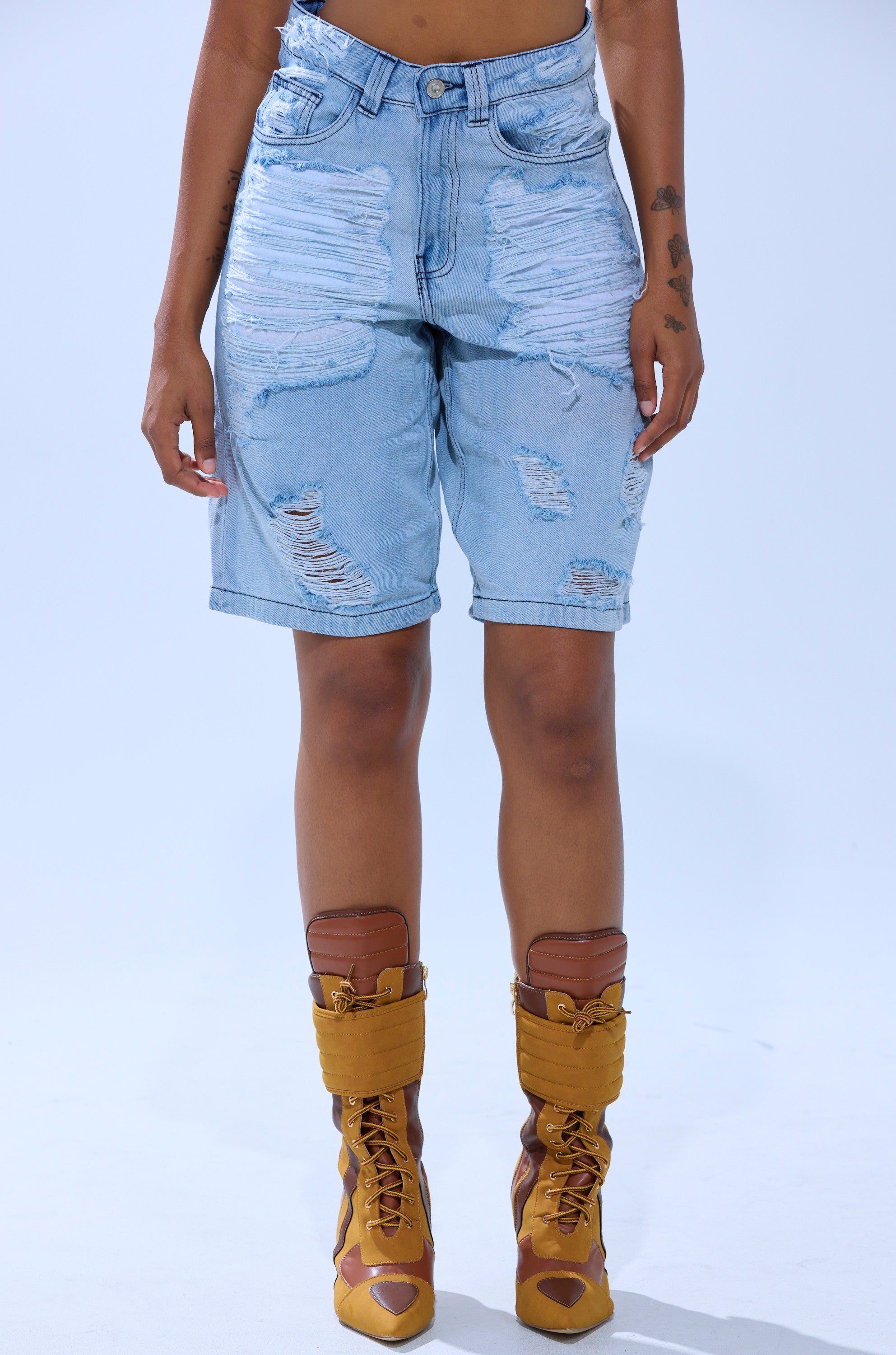 Wearing the KIMMY DISTRESSED BERMUDA SHORT and tan lace-up boots, a person stands against a plain light background, shown from the waist down. 1