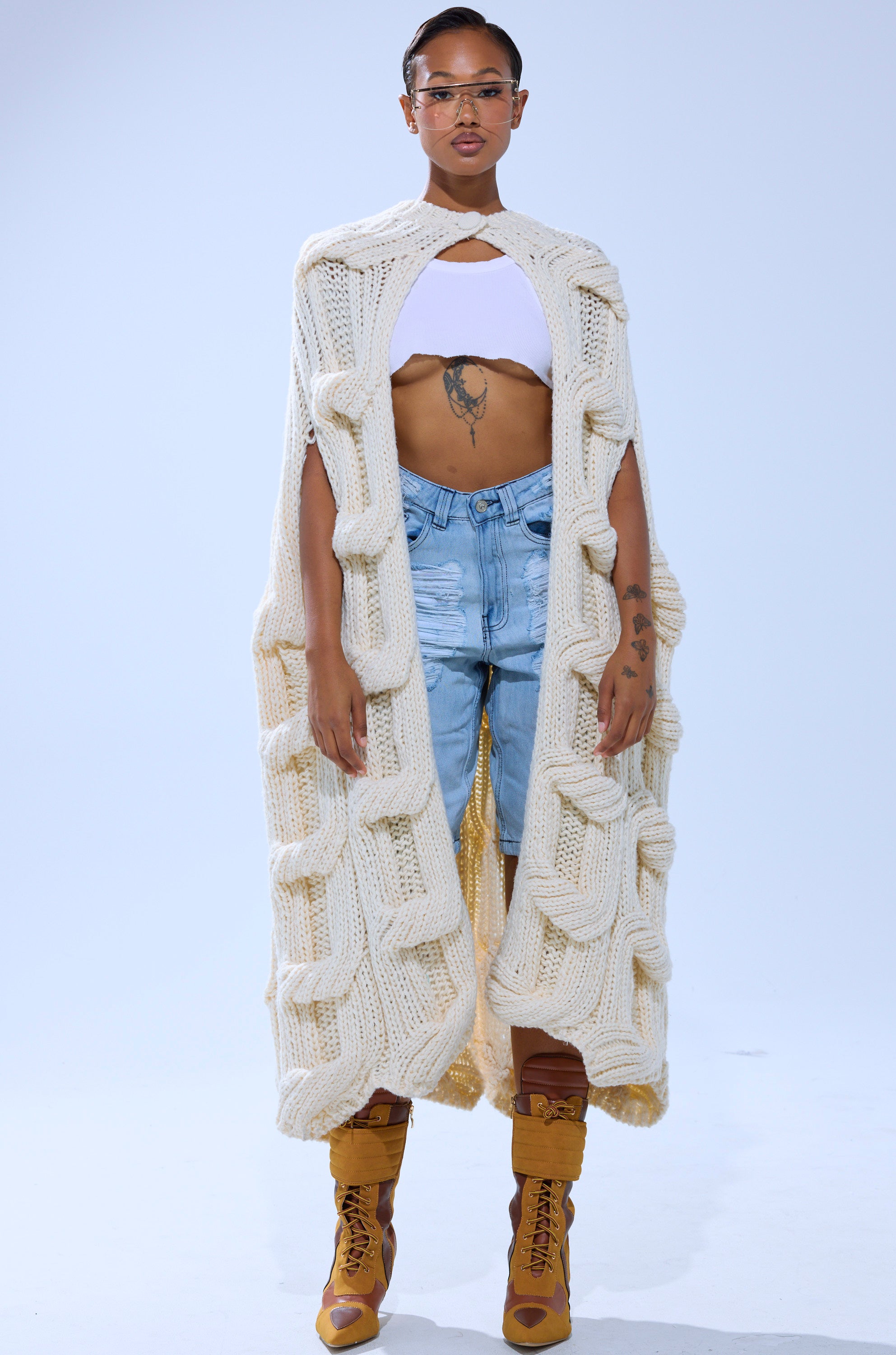 A person stands against a plain background wearing a long, chunky knit cream-colored cape with large textured loops, a white cropped top, KIMMY DISTRESSED BERMUDA SHORTS, yellow heeled boots, and clear glasses. 4