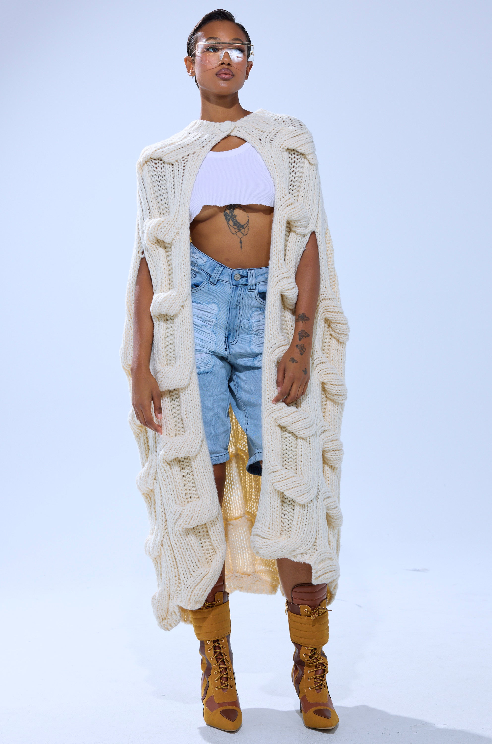 A person in a chunky knit cream cape, white crop top, and KIMMY DISTRESSED BERMUDA SHORT stands against a plain background. Tan lace-up boots, clear glasses, and visible tattoos on midriff and legs add unique details to the look. 5