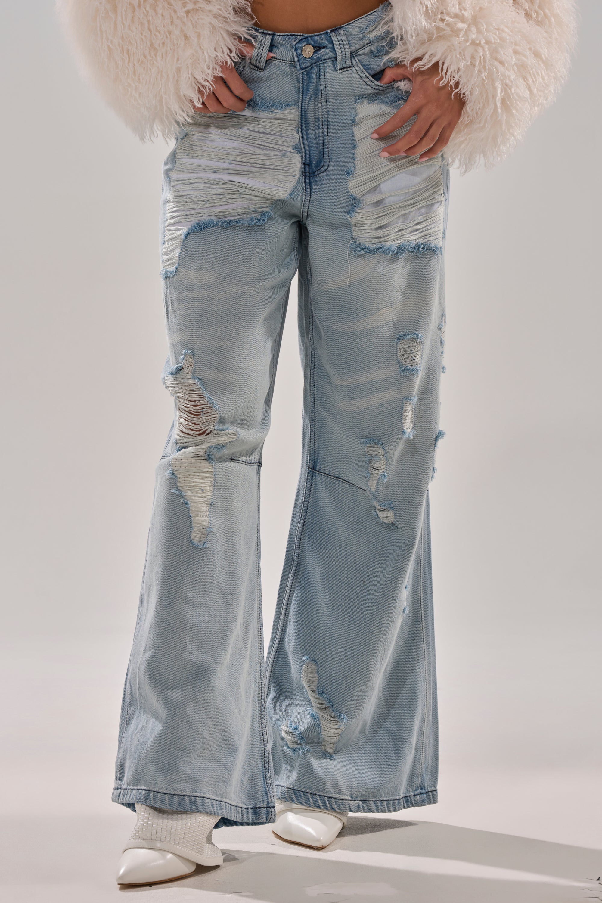 The photo shows someone wearing SUPER DISTRESSED WIDE LEG DENIM—light-wash, ripped, and frayed jeans—paired with white pointed-toe shoes and a fluffy, light jacket. The image is cropped at the torso.
