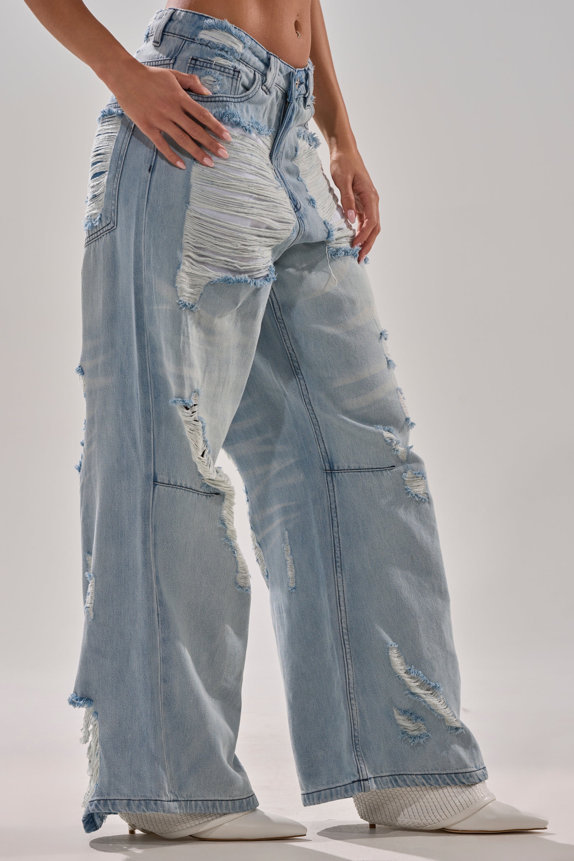 A person models the SUPER DISTRESSED WIDE LEG DENIM, featuring shredded details, paired with white pointed-toe shoes against a plain background.