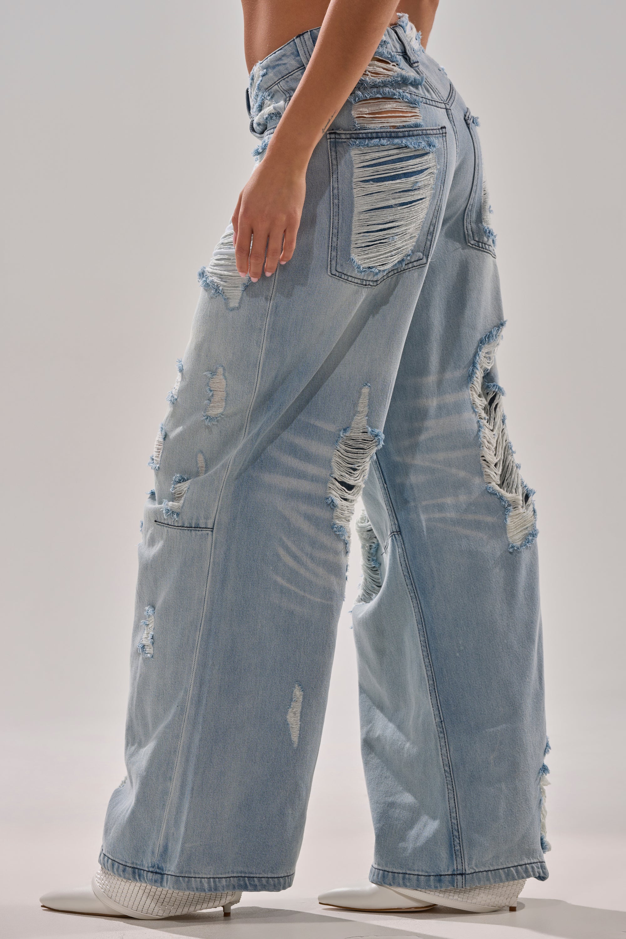 Someone models the SUPER DISTRESSED WIDE LEG DENIM—baggy, light-wash jeans with heavy distressing and shredded details—standing sideways in white heeled shoes against a plain background.