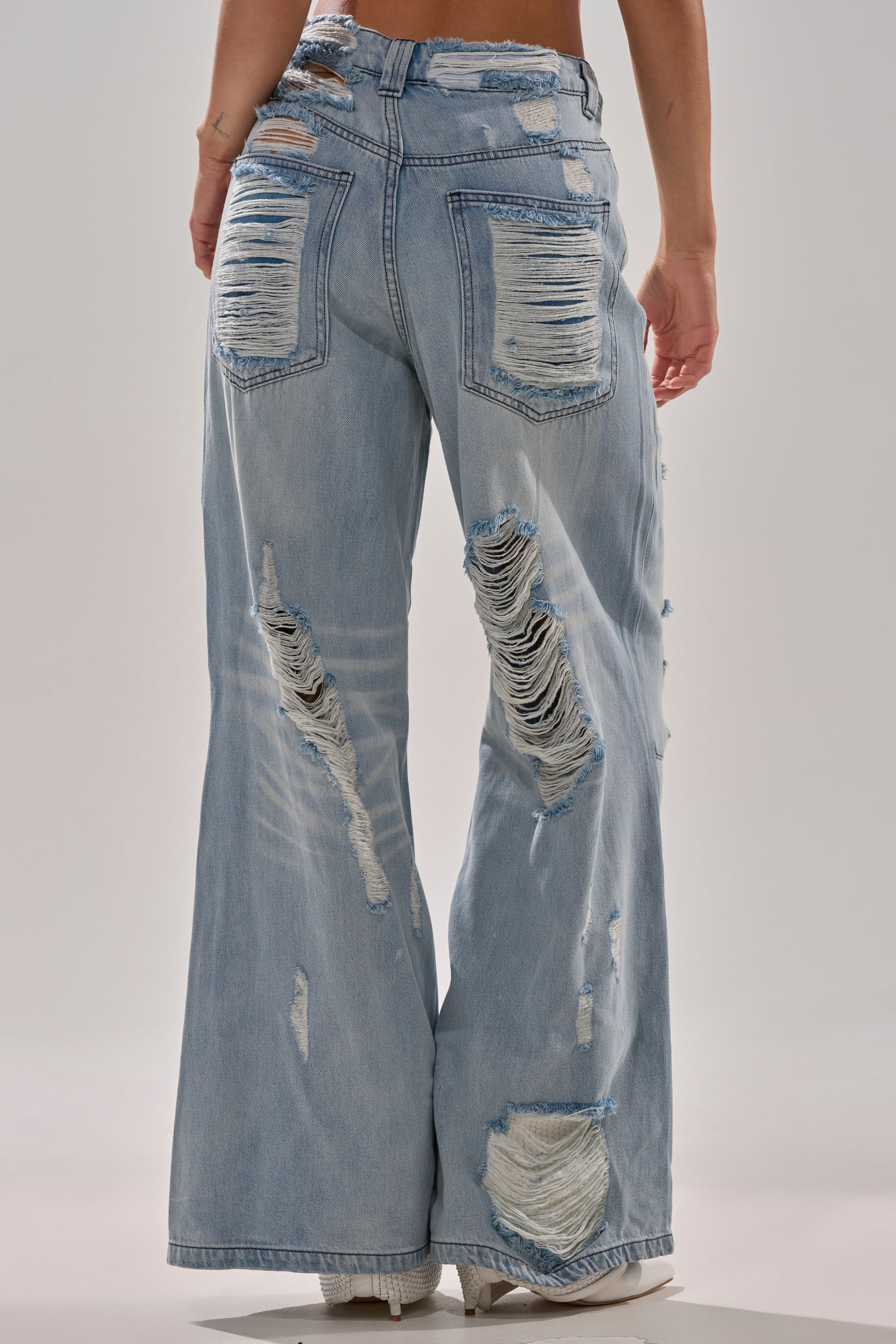 A person wears SUPER DISTRESSED WIDE LEG DENIM, featuring light wash, heavy distressing with large rips on the back pockets, thighs, and calves. The frayed-edge baggy jeans are paired with white shoes.