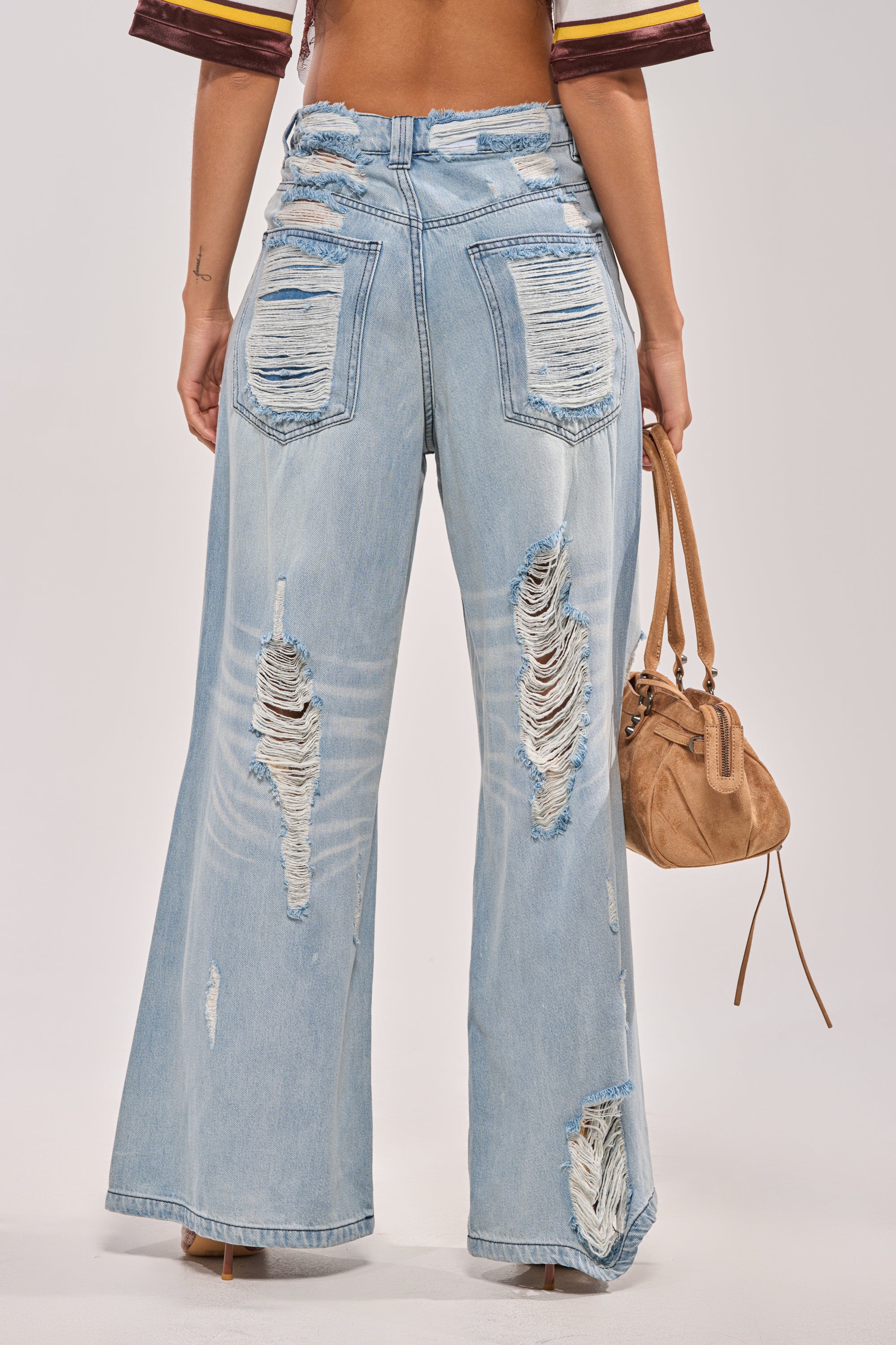 Wearing SUPER DISTRESSED WIDE LEG DENIM with heavy rips on the back, this person holds a small brown suede handbag. Only the lower back and legs of these baggy jeans are shown. 3