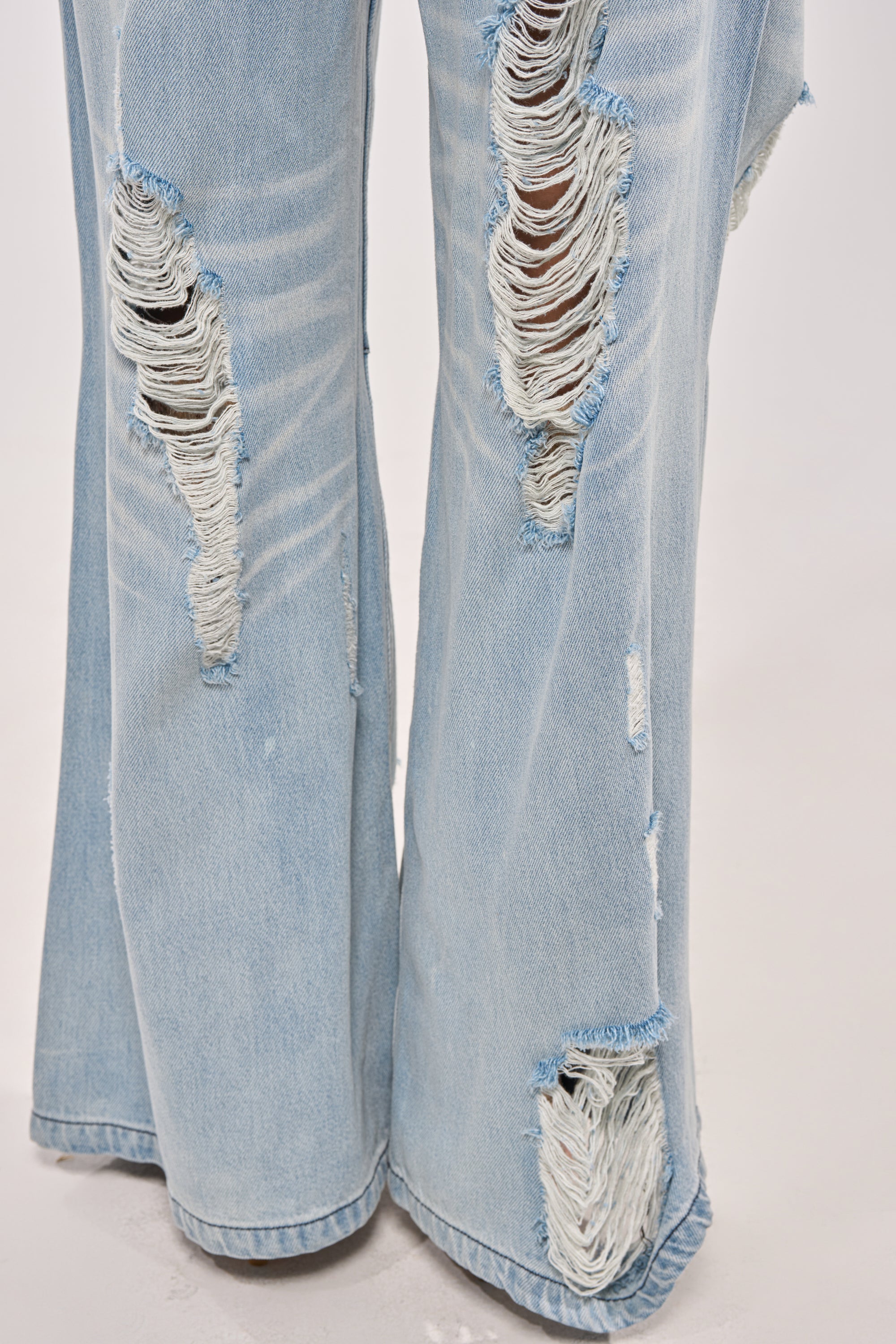 Close-up of lower legs wearing SUPER DISTRESSED WIDE LEG DENIM, featuring light-wash fabric, wide-leg fit, and multiple large rips with frayed details on the front against a plain light background. 6