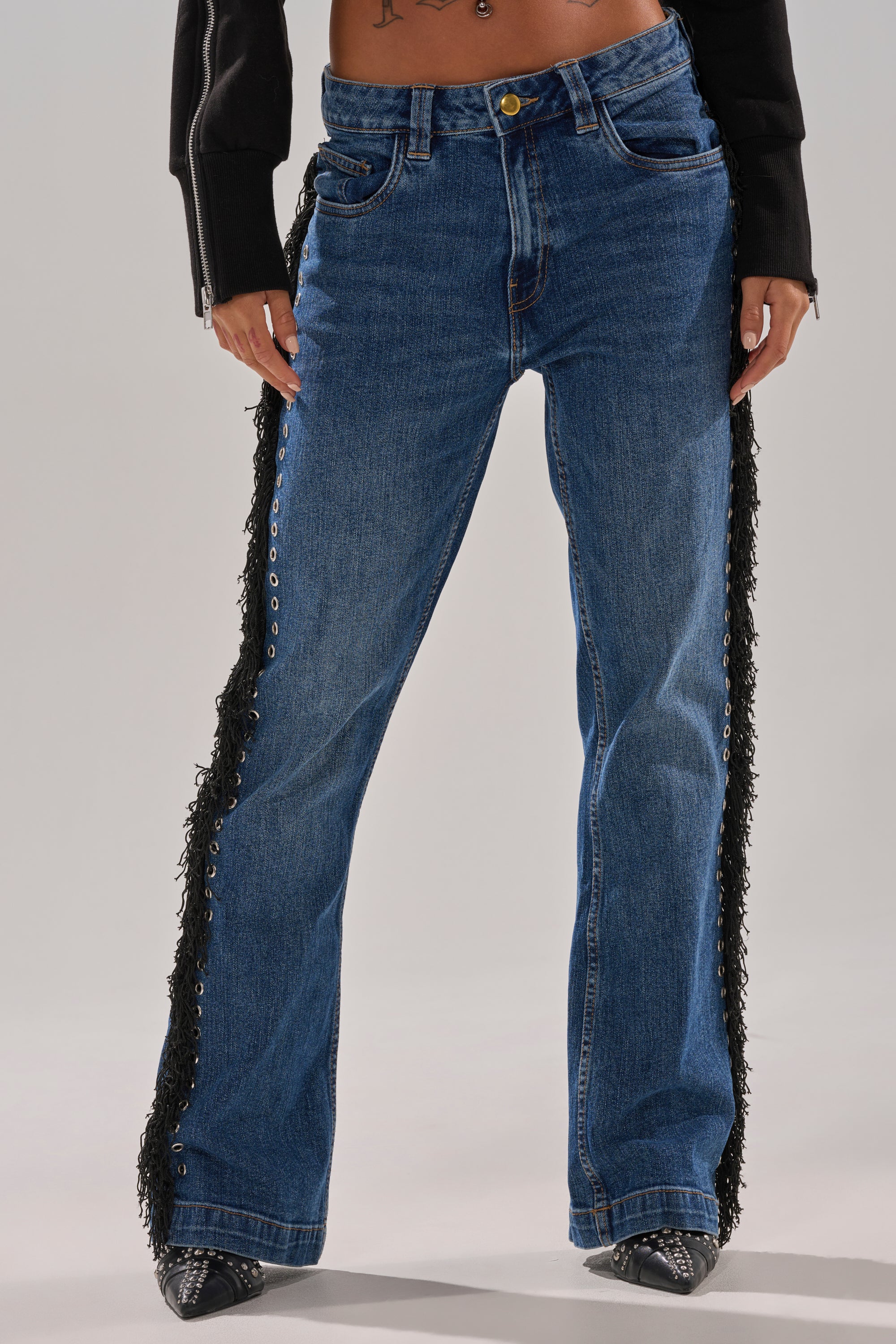 Model wearing PARTY OVER HERE FRINGE STRAIGHT LEG DENIM with black fringe along the outer seams, styled with black pointed-toe shoes and a partially visible black jacket, set against a plain light background.