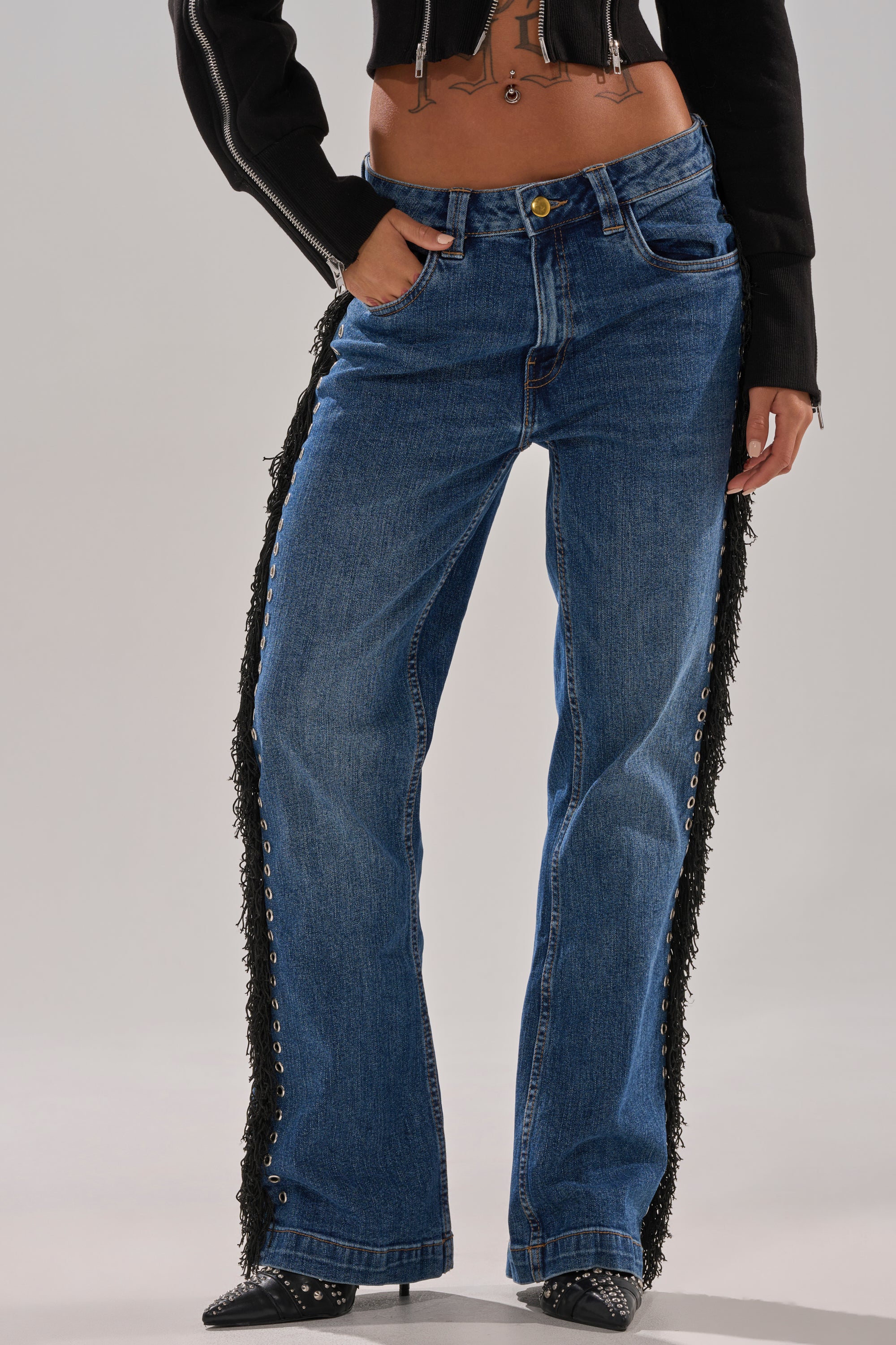 Only the lower half is shown as someone models PARTY OVER HERE FRINGE STRAIGHT LEG DENIM—blue jeans with black fringe and grommets—styled with a cropped black jacket and pointed black boots.
