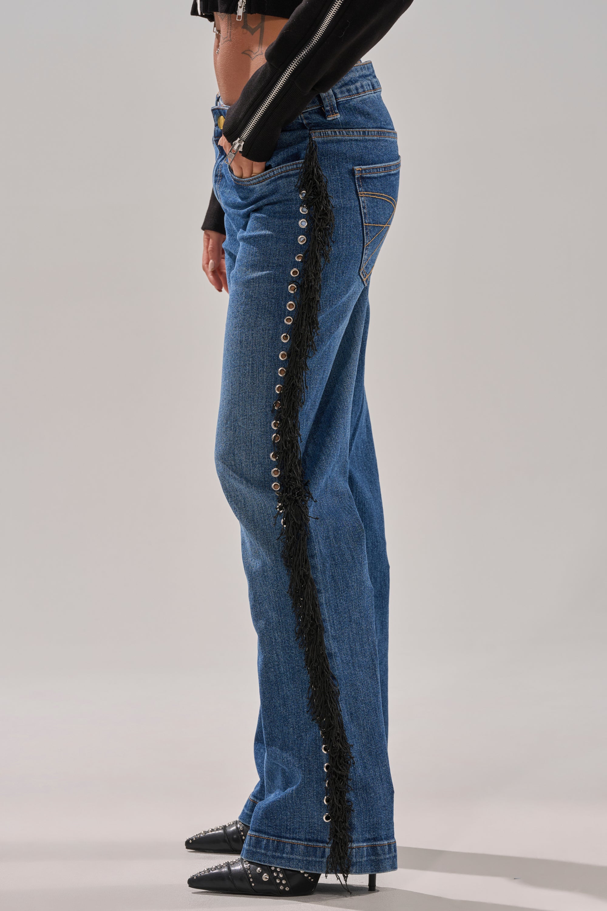 A person wearing the PARTY OVER HERE FRINGE STRAIGHT LEG DENIM with black metal eyelets, styled with a black cropped top and pointed studded shoes—bold statement party jeans. Photo shows a side view.