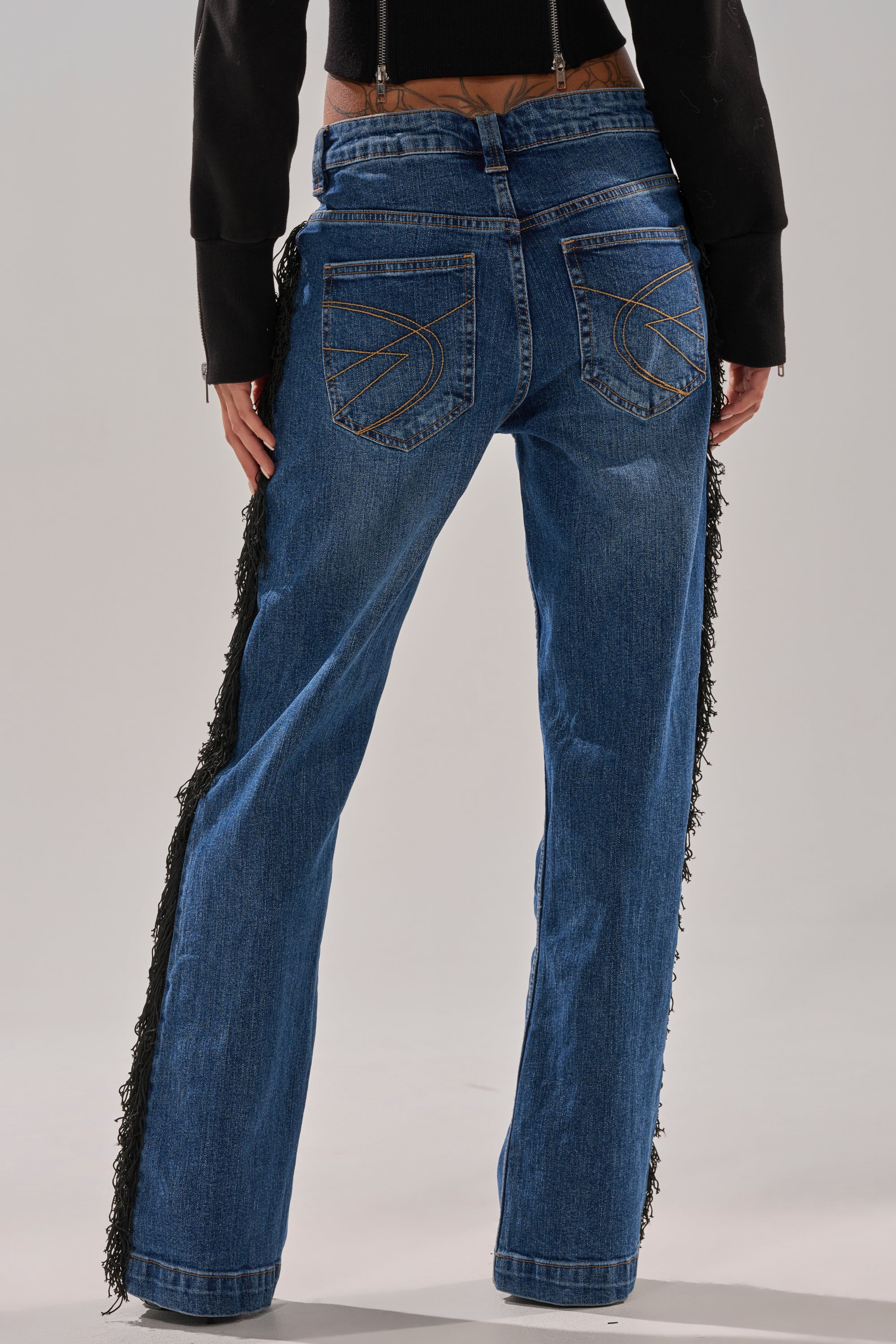 A person is shown from the back wearing the PARTY OVER HERE FRINGE STRAIGHT LEG DENIM—blue jeans with decorative pocket stitching and black fringe down the sides—paired with a black jacket.