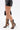 Wearing a black faux leather mini skirt, a person stands against a plain white background in AZALEA WANG INDRANI BLACK GLADIATOR SANDALS with knee-high, snakeskin embossing and stiletto heels, holding a black handbag.