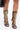A close-up shows a person wearing AZALEA WANG INDRANI BLACK GLADIATOR SANDALS with knee-high stiletto heels, bold snakeskin embossing, and black trim, paired with a shiny black handbag against a plain white background.