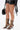 A person stands against a white background wearing black faux-leather shorts, carrying a black handbag, and sporting the AZALEA WANG INDRANI BLACK GLADIATOR SANDAL.