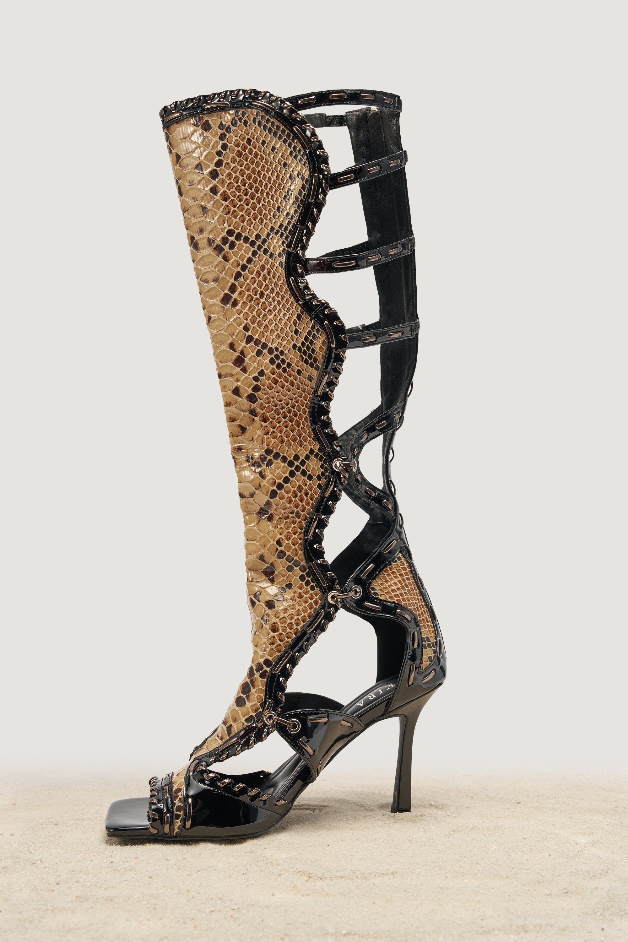 The AKIRA GOLD INDRANI BLACK HIGH SHAFT GLADIATOR SANDAL features knee-high black patent leather straps, an open toe, stiletto heel, and a snakeskin pattern, showcased against a neutral background. 3